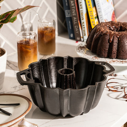 A black seasoned cast iron fluted cake pan is displayed on a table with a baked chocolate bundt cake in the background, along with some books and a glass of beverage.