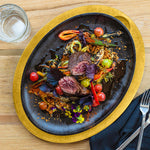 Delicate dish of beef with vegetables on a cast iron serving griddle with a yellow underliner, served on a kitchen table.