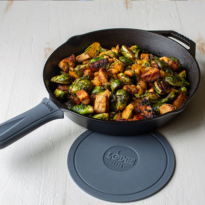 Grey silicone handle holder on skillet with freshly made chicken and Brussel sprouts next to Lodge deluxe silicone trivet.