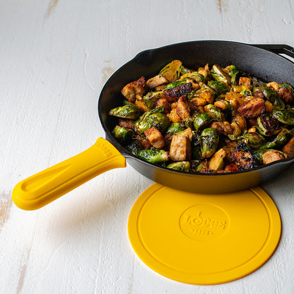Yellow silicone handle holder on skillet with freshly made chicken and Brussel sprouts next to Lodge deluxe silicone trivet.