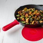 Red silicone handle holder on skillet with freshly made chicken and Brussel sprouts next to Lodge deluxe silicone trivet.