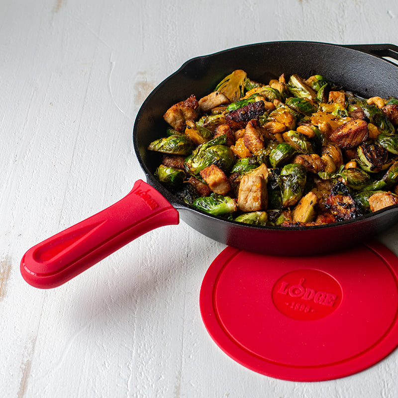 Red silicone handle holder on skillet with freshly made chicken and Brussel sprouts next to Lodge deluxe silicone trivet.