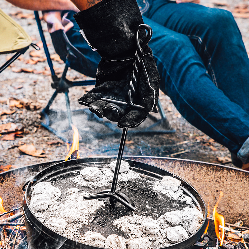 A person is using a black lid lifter to lift the lid of a camp dutch oven placed on a campfire.