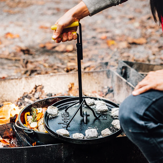 A person is using a black lid lifter with a yellow grip to lift the lid of a camp dutch oven placed on a campfire.