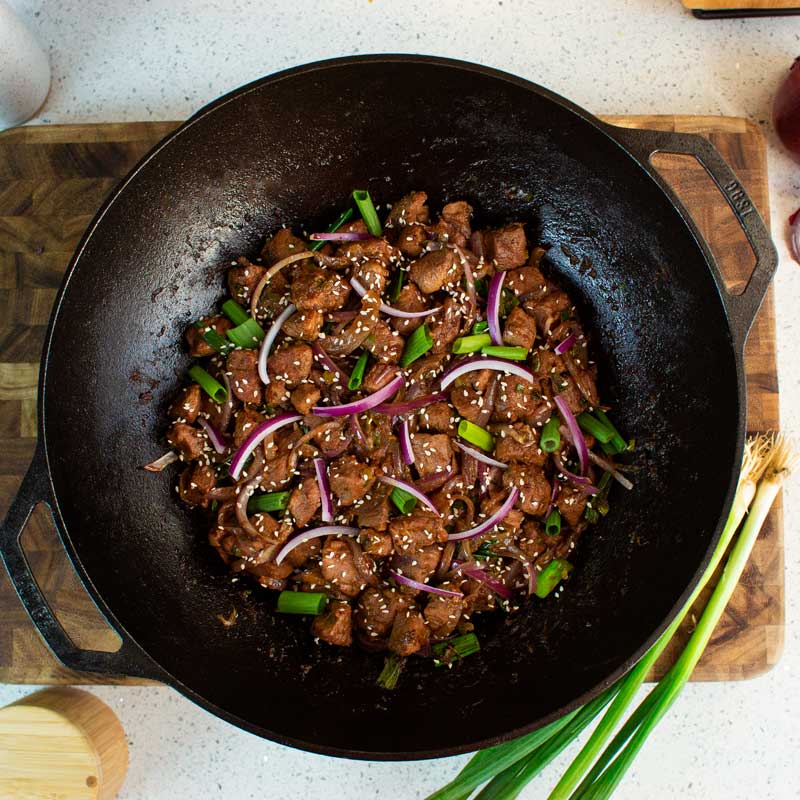 Fried beef dish with onions and green onions in a cast iron wok on a wooden cutting board.