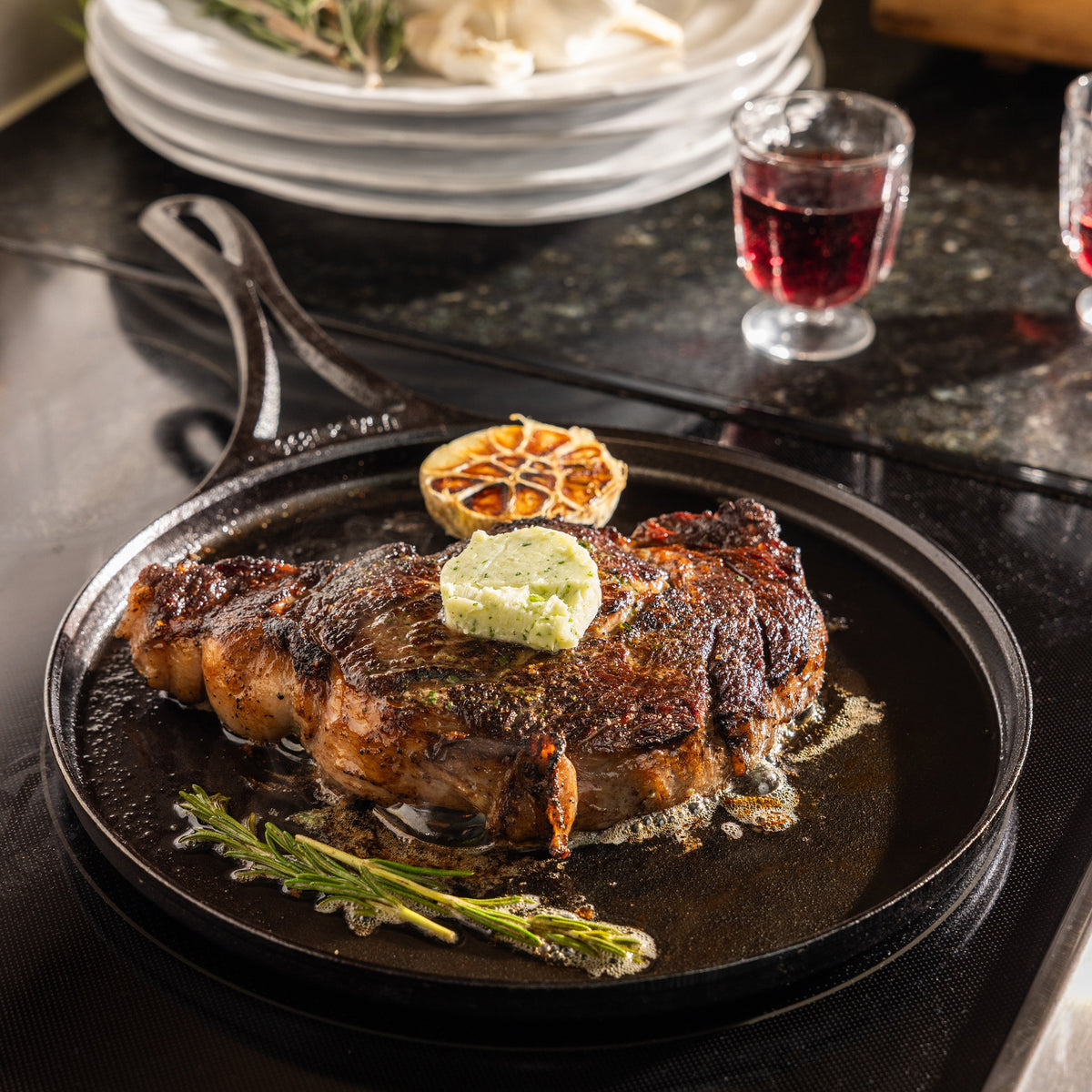 A seared steak topped with a dollop of herb butter and a halved lemon, served in a black cast-iron skillet on a dark tabletop with plates and a glass of red wine in the background.