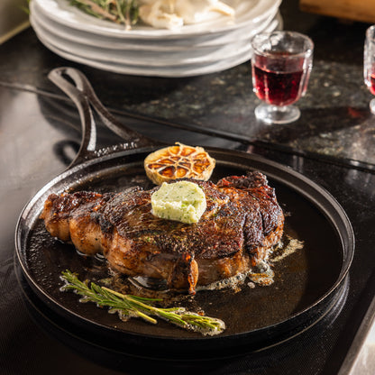 A seared steak topped with a dollop of herb butter and a halved lemon, served in a black cast-iron skillet on a dark tabletop with plates and a glass of red wine in the background.