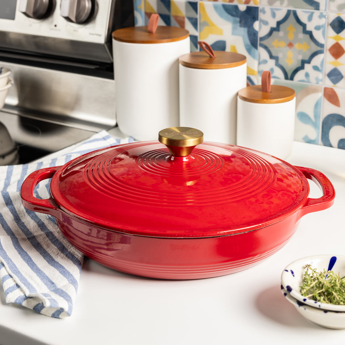Red casserole dish with gold lid knob on a kitchen counter, with canisters and a colorful backsplash.