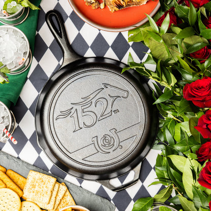 Seasoned Cast Iron Kentucky Derby Skillet for the 152nd year, placed on checkered table surrounded by food and roses.