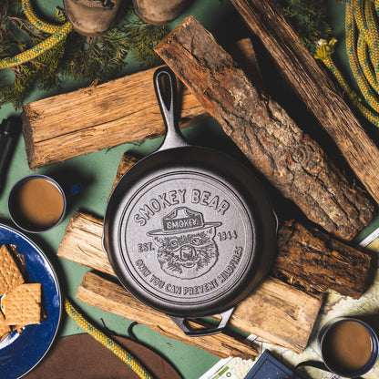Cast iron skillet with 'Smokey Bear' branding on a rustic setting with logs and coffee cups.