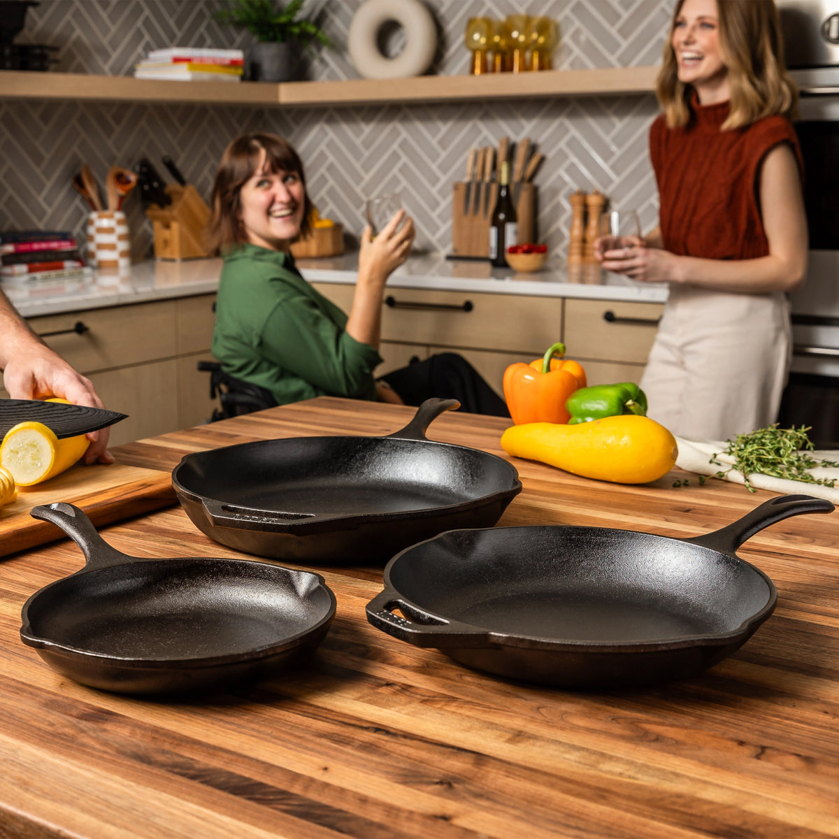 Three cast iron skillets on a wooden countertop with two women in a kitchen.