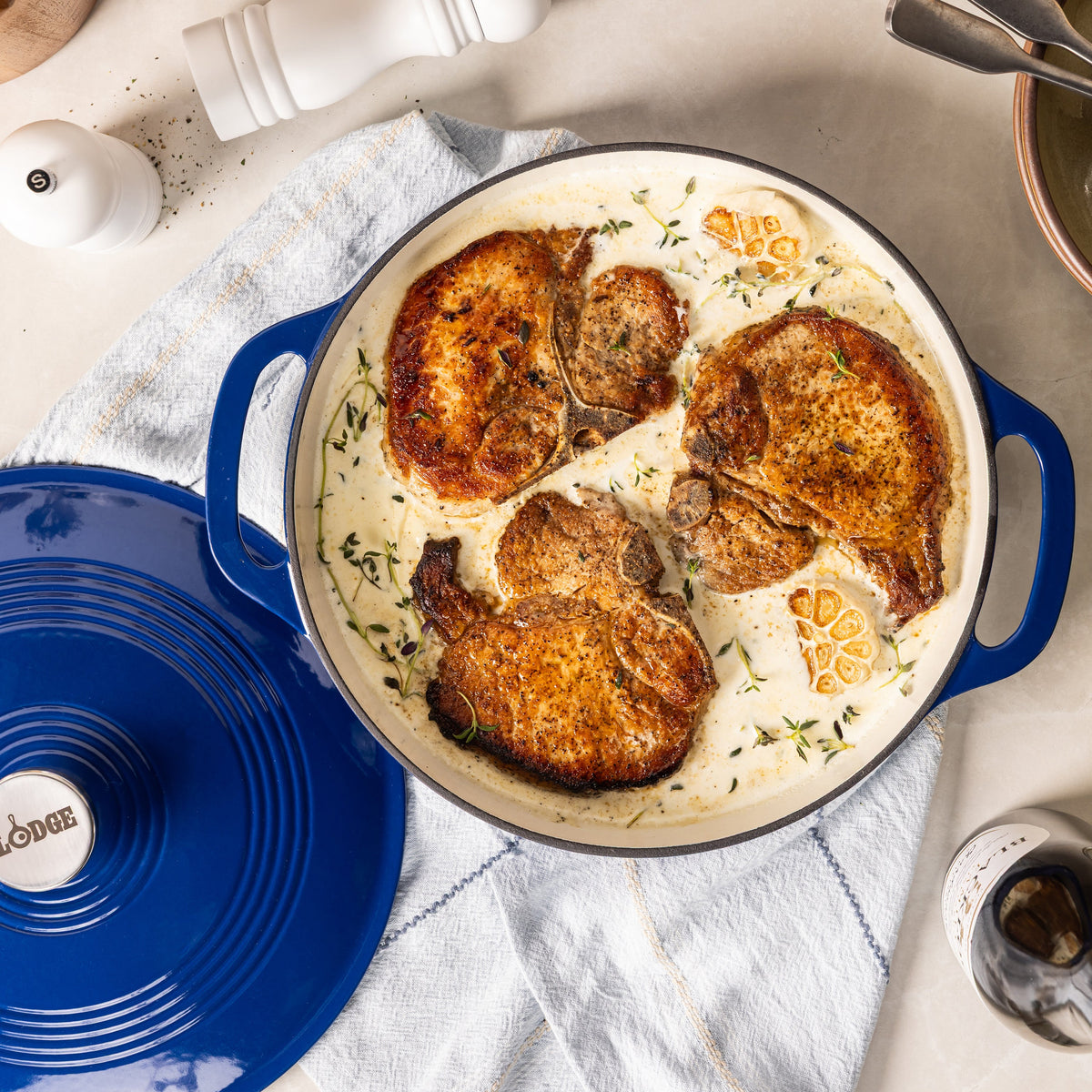 A top-down view of a blue enameled cast iron braiser containing cooked chicken and cream sauce with lemon slices, alongside the matching blue lid on a light countertop.