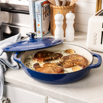 A blue enameled cast iron oval casserole dish with a matching lid sits on a kitchen counter, containing cooked chicken breasts in a creamy sauce. The background shows a kitchen setting with a cookbook, salt and pepper shakers, and a toaster.