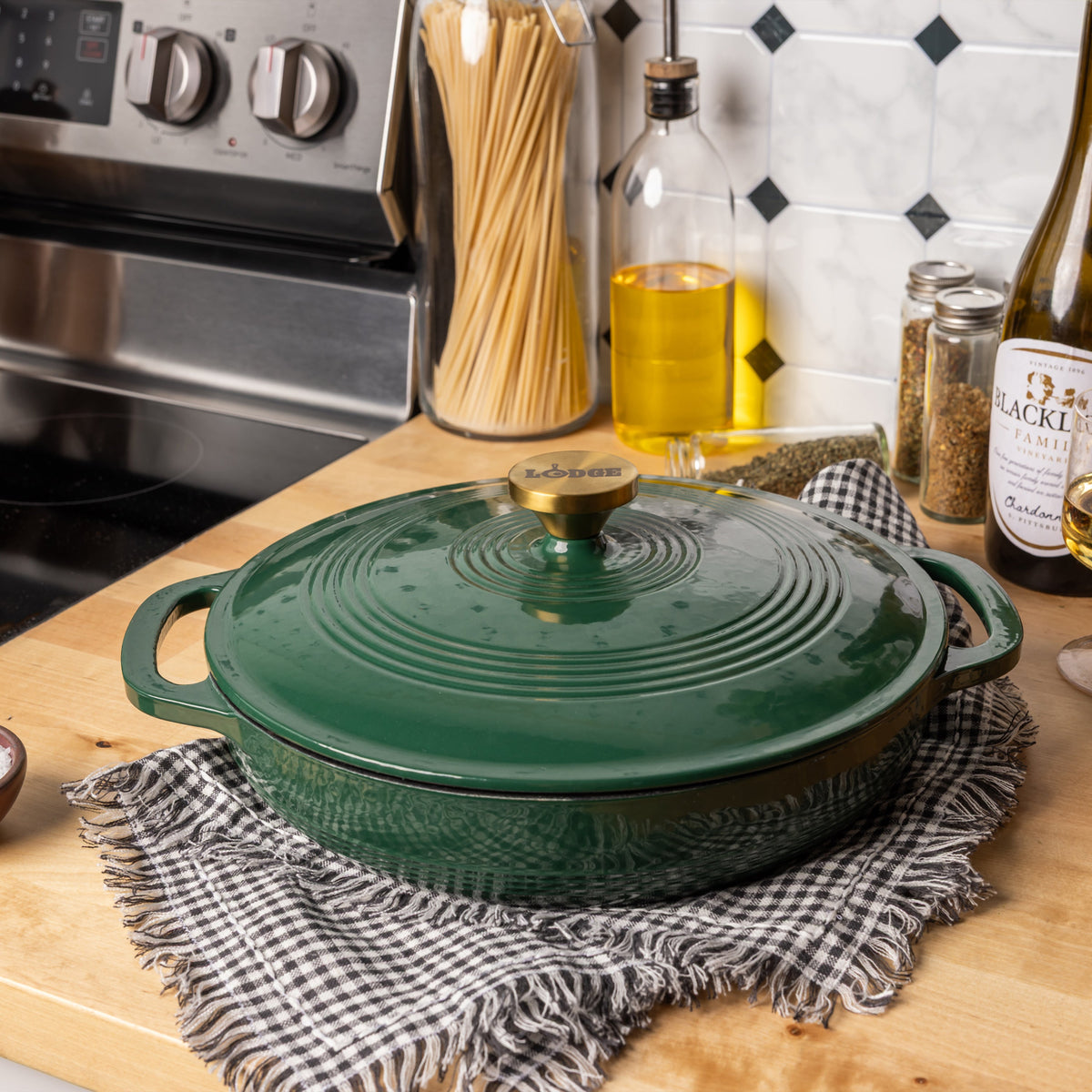 A dark green enameled cast iron braiser with a matching lid and brass knob, resting on a black-and-white checkered cloth on a wooden countertop in a kitchen setting.