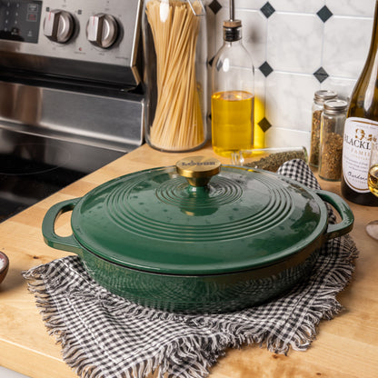 A dark green enameled cast iron braiser with a matching lid and brass knob, resting on a black-and-white checkered cloth on a wooden countertop in a kitchen setting.