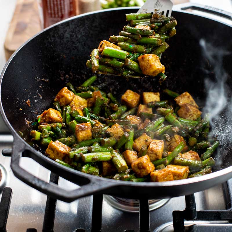 Someone turning tofu and asparagus in wok while cooking on stove.
