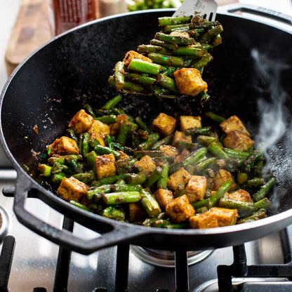 Someone turning tofu and asparagus in wok while cooking on stove.