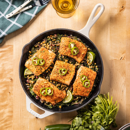 A cast iron skillet with white handles sits on a wooden table, filled with seasoned fish fillets topped with sliced jalapeño and lime, served over a bed of greens and nuts. A glass of beer, a folded napkin, and fresh cilantro are arranged around the skillet.