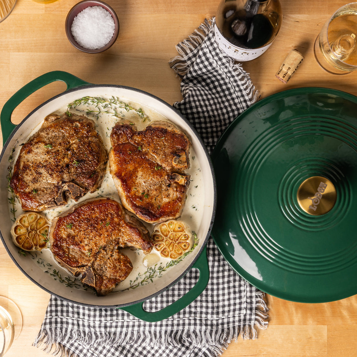 A green enameled cast iron braiser with three cooked pork chops, lemon slices, and herbs, shown alongside its matching green lid on a wooden table with dining props.