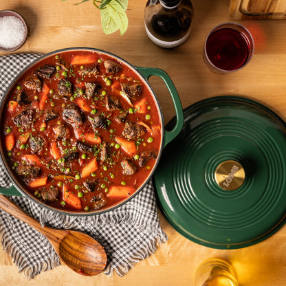 A green enameled cast iron Dutch oven filled with a hearty beef and carrot stew, shown on a wooden table alongside its matching lid, a wooden spoon, a checkered cloth, and dining props.
