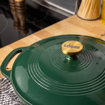 A close-up of a dark green Lodge enameled cast iron pot with a matching lid and a gold-tone knob, sitting on a wooden countertop in a kitchen.