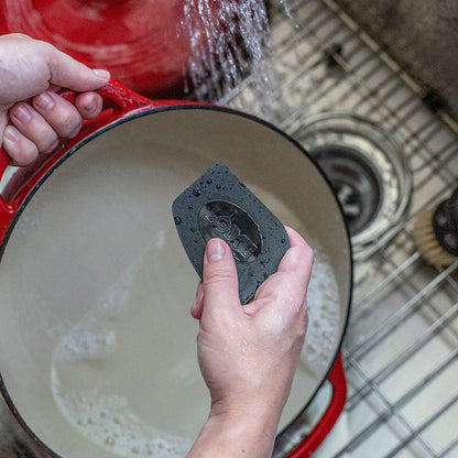 Black scraper being using in kitchen sink to clean enameled dutch oven.