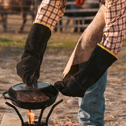 Someone using leather cloves while cooking with a skillet and cast iron grill press over a fire.