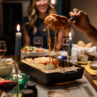 Person lifting a slice of lasagna with stringy cheese from a pan on a dinner table.
