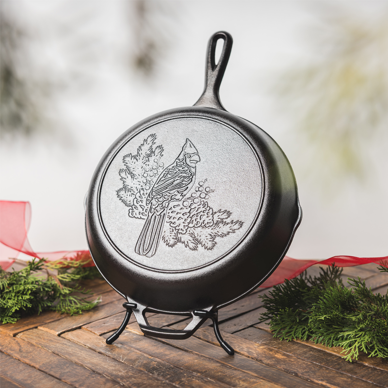Cast iron skillet with a cardinal engraving, displayed on a Lodge Skillet Display stand with a festive holiday background.