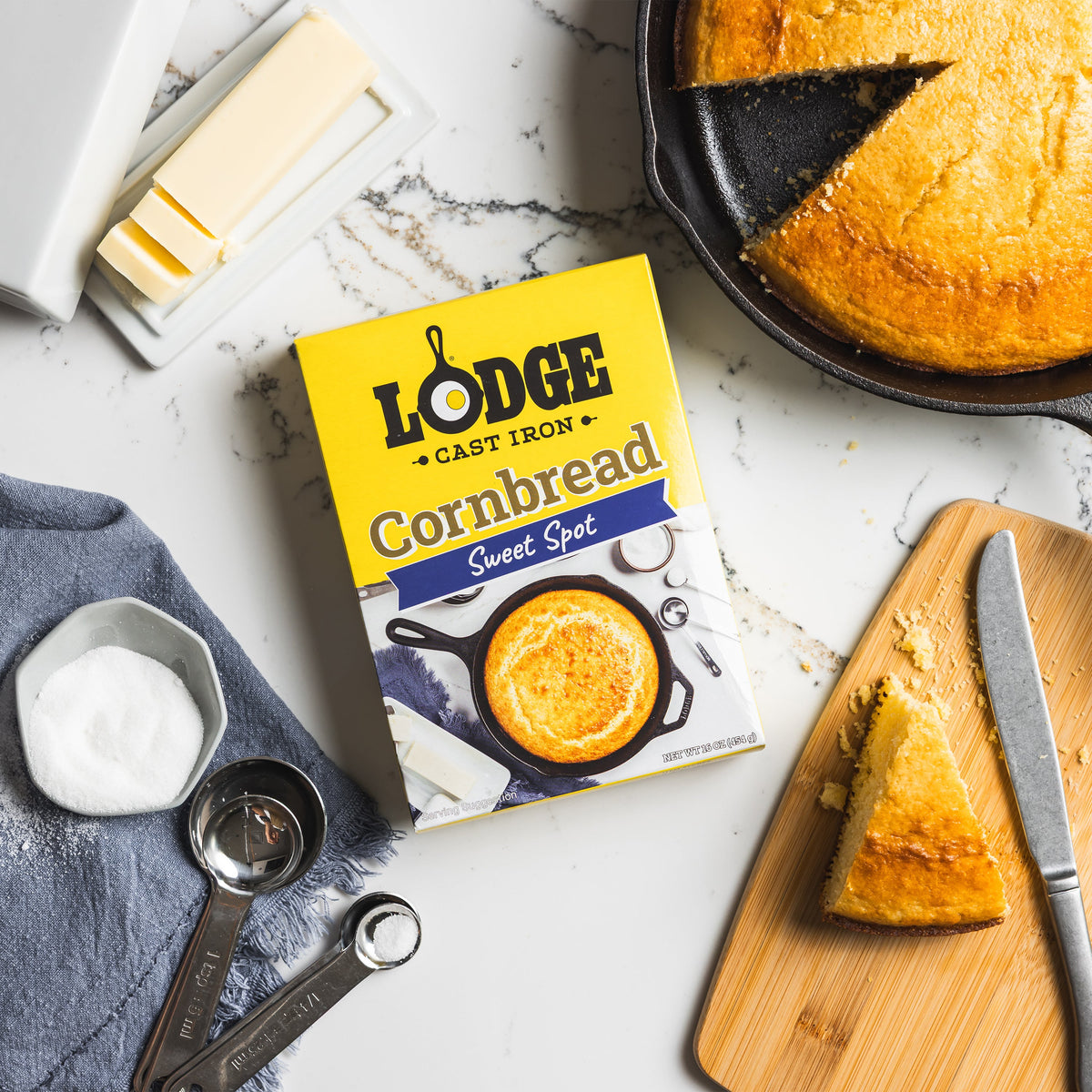 Lodge cornbread mix box with cornbread on a cutting board, knife, and measuring cups on a marble surface.