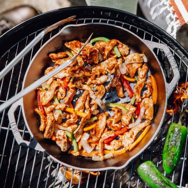 Someone making chicken fajitas with grill basket on charcoal grill while roasting peppers nearby.