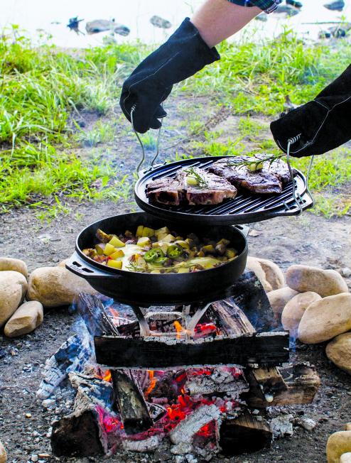 Someone using the hook and carry handles to lift grill pan/griddle after searing steak. Below the grill pan is the wok portion cooking a stew over the fire using our Fire and Cook Stand.
