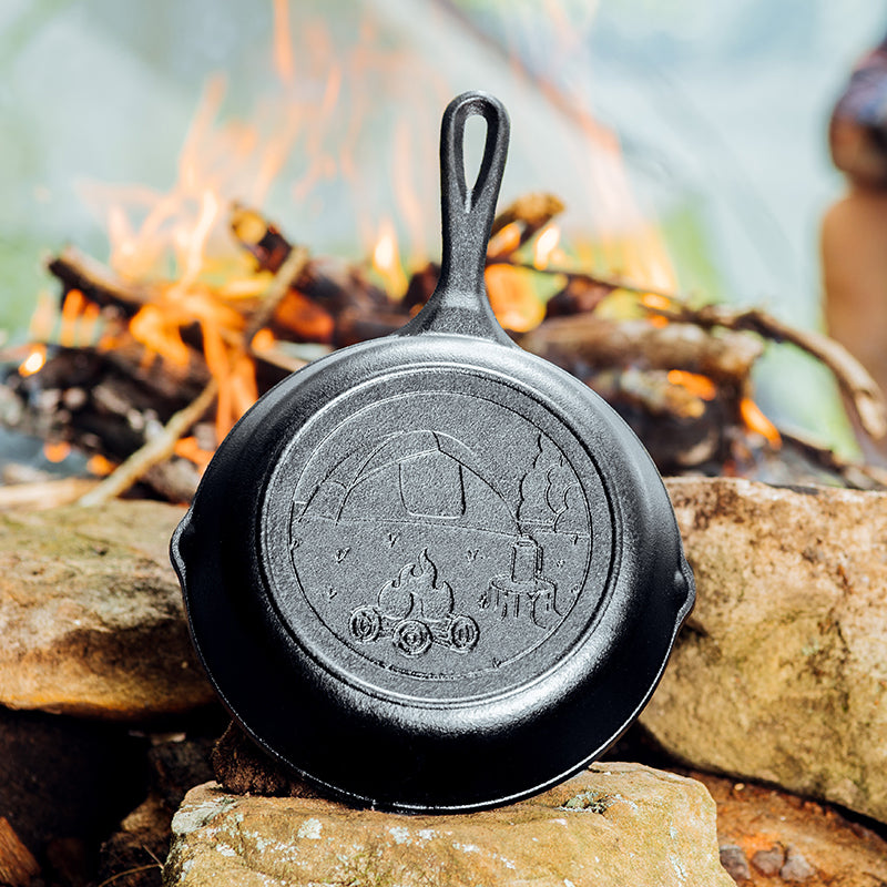 A black cast iron skillet with engraved campsite design, placed on a stone surface with a camping fire in the background.