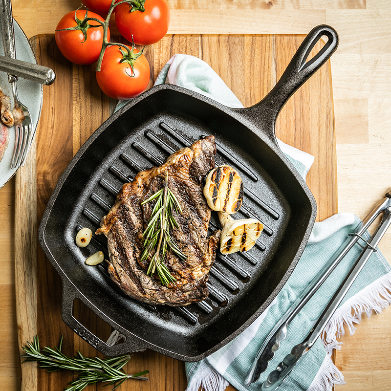 Freshly cooked steak garnished with garlic and rosemary, presented in grill pan on table.