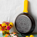 Decorative frying pan with skull design on a white wall with flowers and a cake.