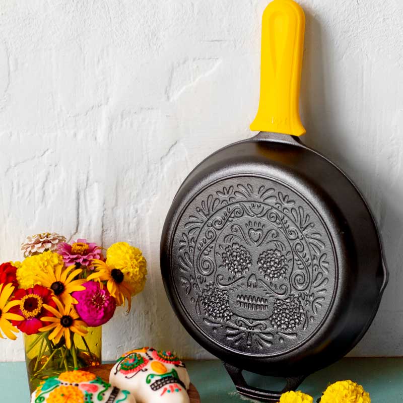 Decorative frying pan with skull design on a white wall with flowers and a cake.