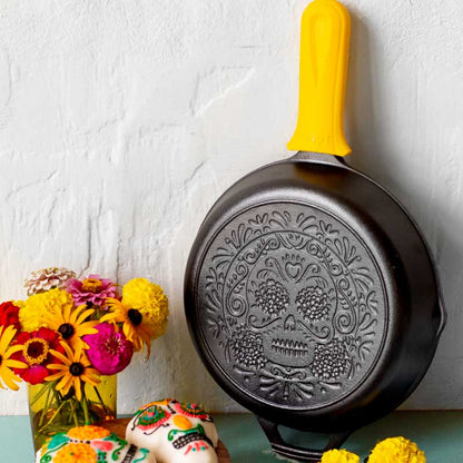 Decorative frying pan with skull design on a white wall with flowers and a cake.