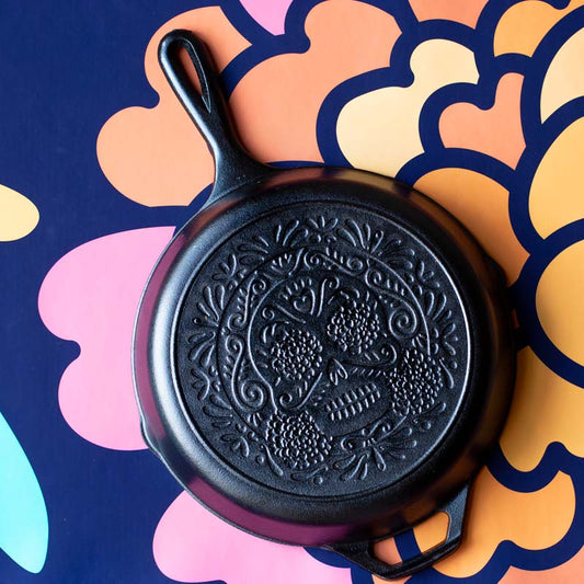 A black cast iron skillet with a sugar skull pattern, displayed on a colorful background with floral motifs.