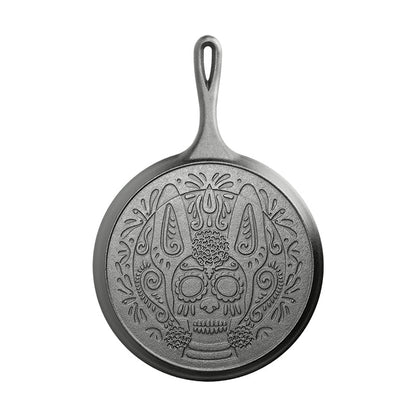 The back of a 10.5 inch round seasoned cast iron griddle with a handle, featuring a sugar skull design resembling a Xolo dog.