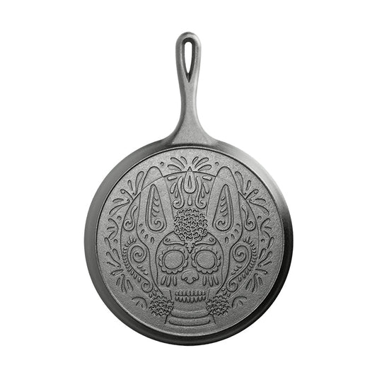 The back of a 10.5 inch round seasoned cast iron griddle with a handle, featuring a sugar skull design resembling a Xolo dog.