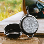 A black cast iron cabin combo cooker, complete with 2 pieces, one being a skillet lid, featuring a engraved cabin design, and the other being a deep skillet. Both pieces are displayed at a campsite.