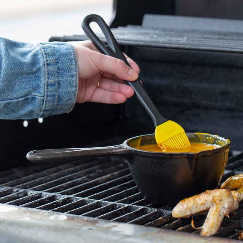 Someone using a basting brush to mix a sauce in pot over grill.
