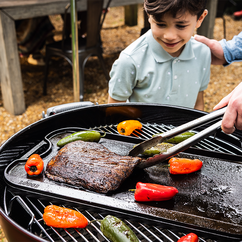Someone turning seared steak on griddle on a charcoal grill while roasting peppers.