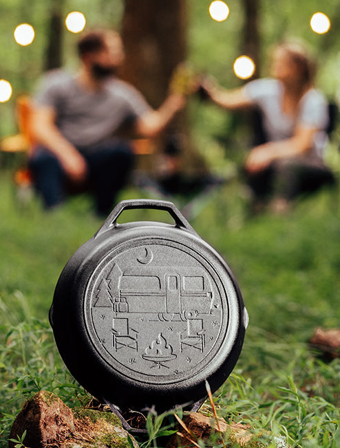 A cast iron camping pan with a dual handle design, featuring an engraved camper scene, placed on the grass with a blurred background of a couple sitting in the distance.