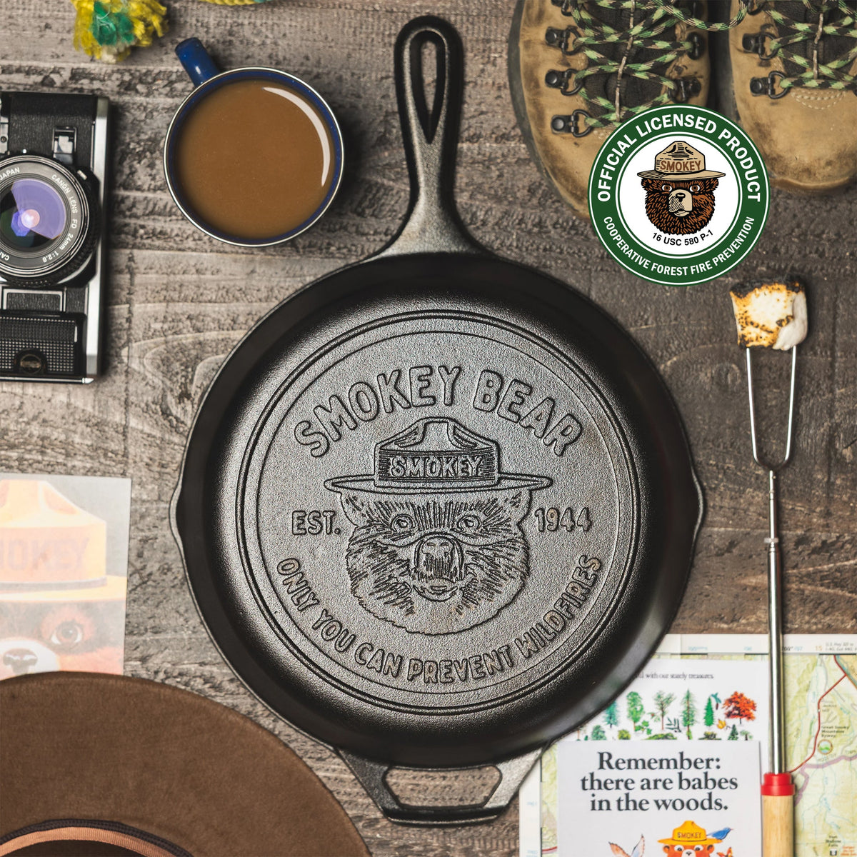 Cast iron skillet with Smokey Bear logo on a rustic wooden surface with coffee, boots, and a camera.