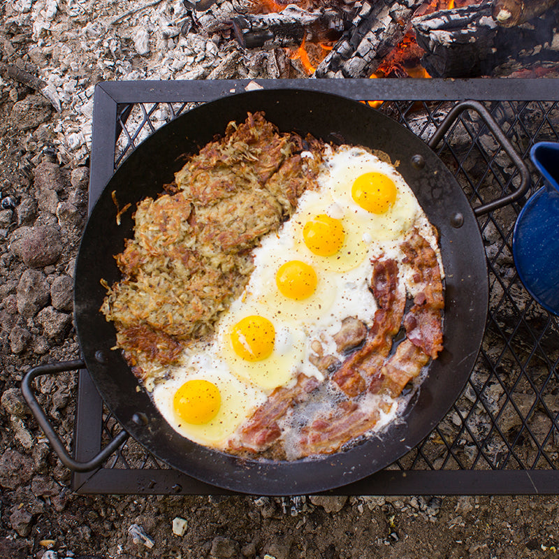 A full breakfast of eggs, hashbrowns, and bacon being cooked in pan near campfire.