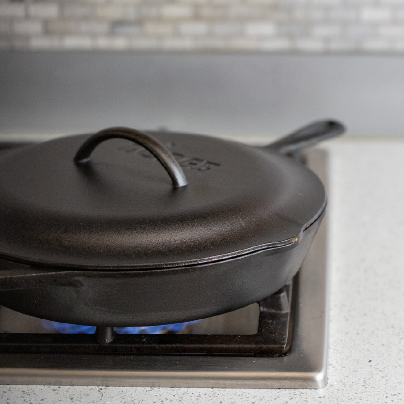 A black cast iron lid placed on top of a cast iron skillet on a stovetop, indicating use for roasting, braising, and simmering.