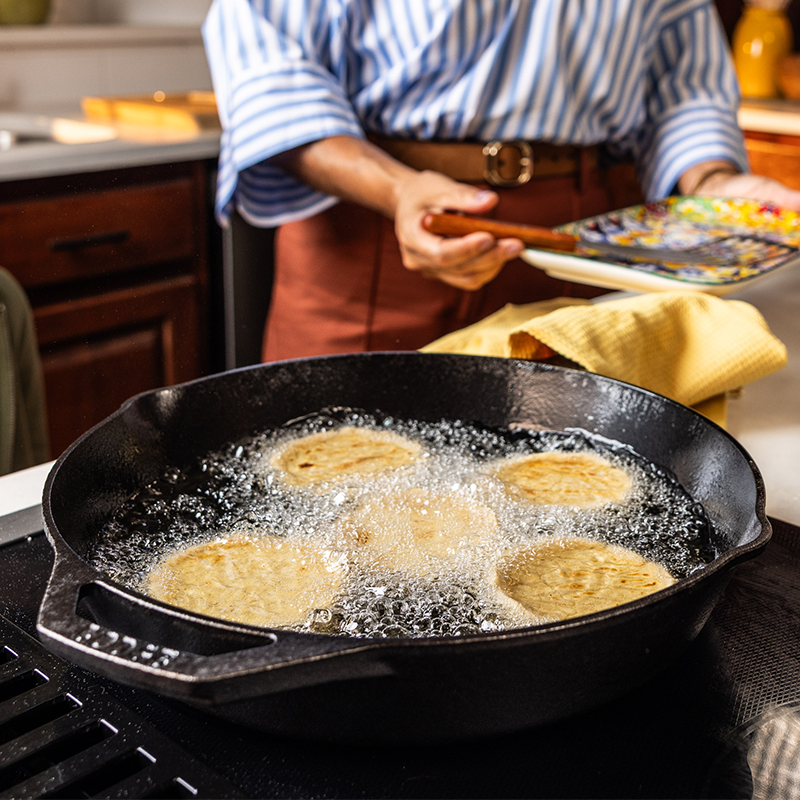 Person frying tortillas in a cast iron skillet with a kitchen setting in the background.
