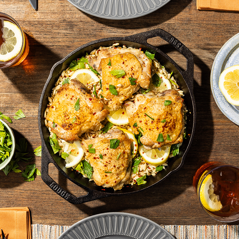 Skillet of chicken with lemon and herbs on a wooden table with drinks and plates.