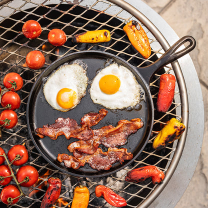 Bacon and eggs cooking on griddle over charcoal grill while peppers and tomato's roast around it..
